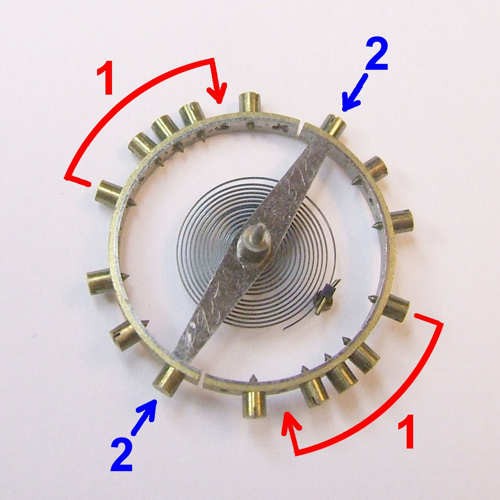 Screwed bimetallic balance wheel from an old pocket watch with its spiral hairspring, on a white background, with red and blue arrows marking parts
