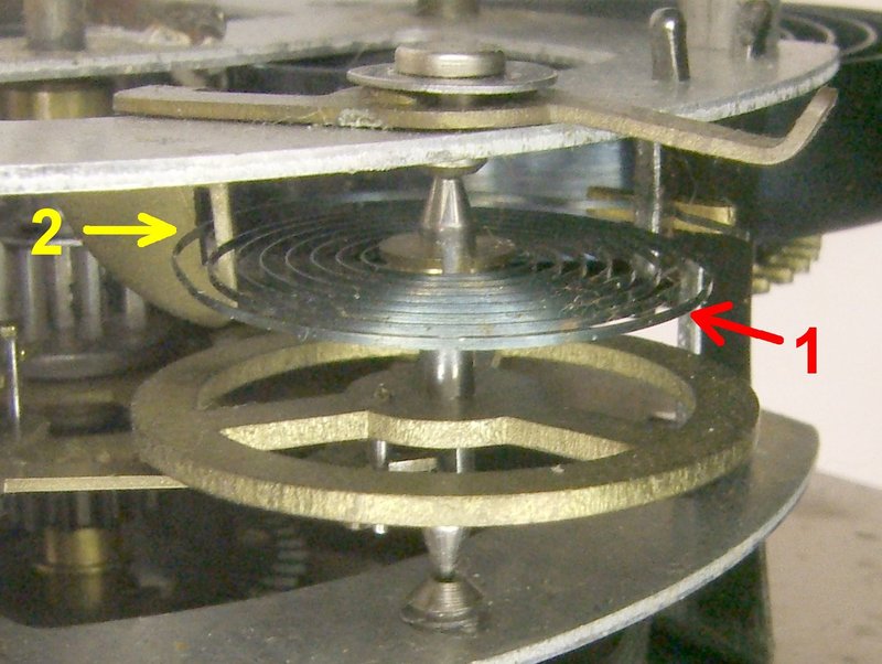 Balance wheel inside a 1950s alarm clock with yellow and red arrows pointing at the hairspring and the regulator