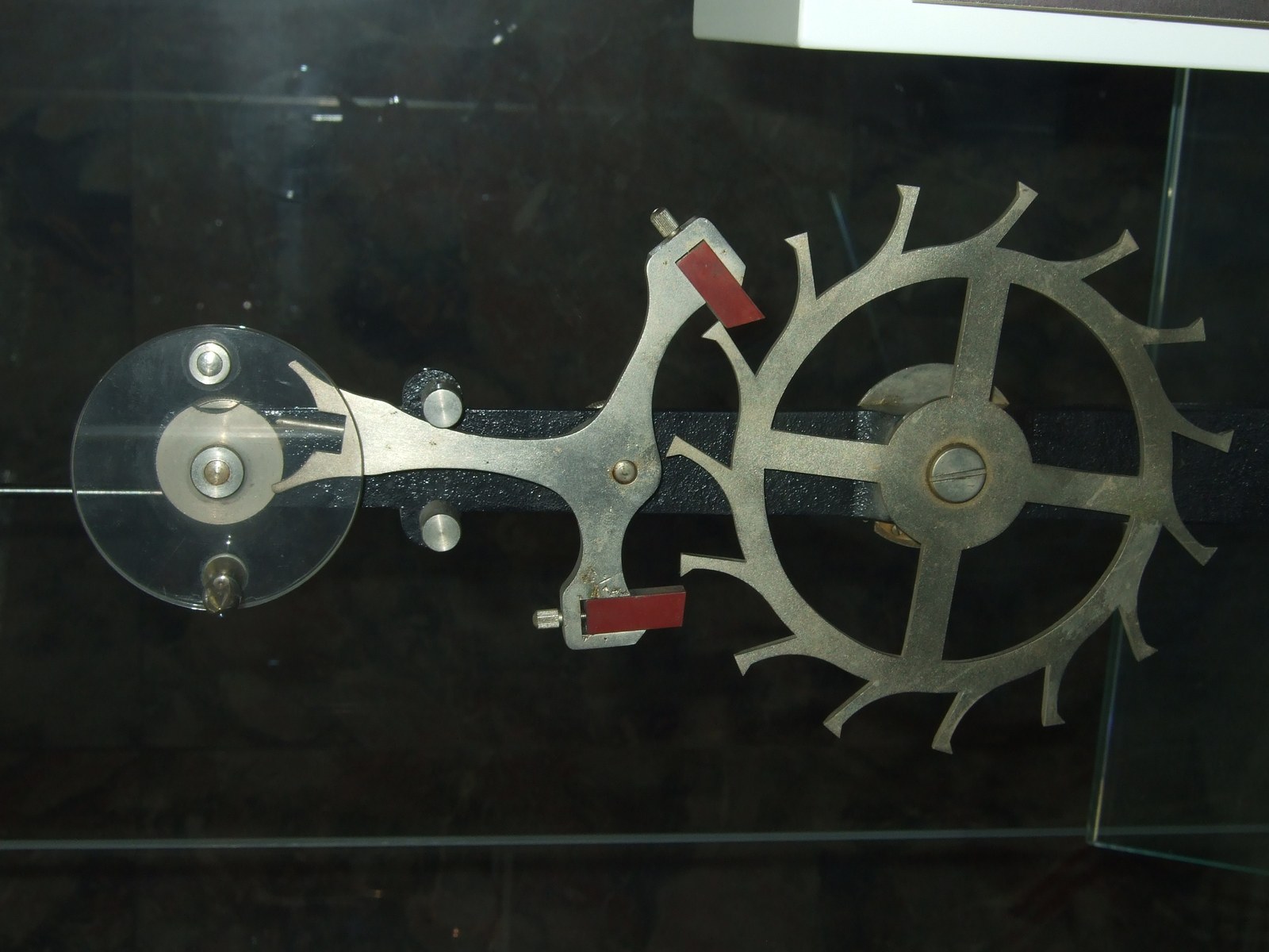 Large working model of a lever escapement on a dark background, with a clear balance, a steel pallet lever with red pallet stones, and an escape wheel